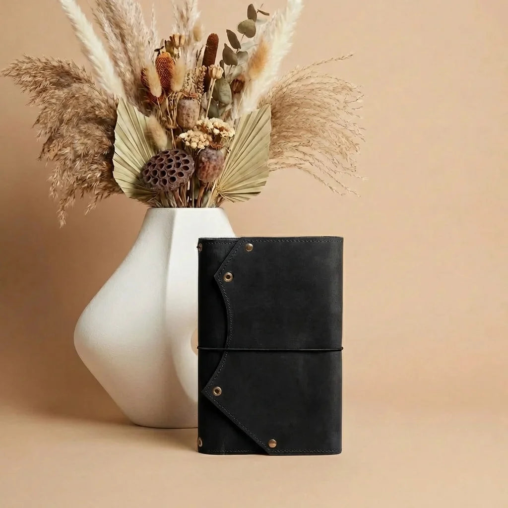 Black leather clutch with gold accents next to a white vase with dried plants on a beige background