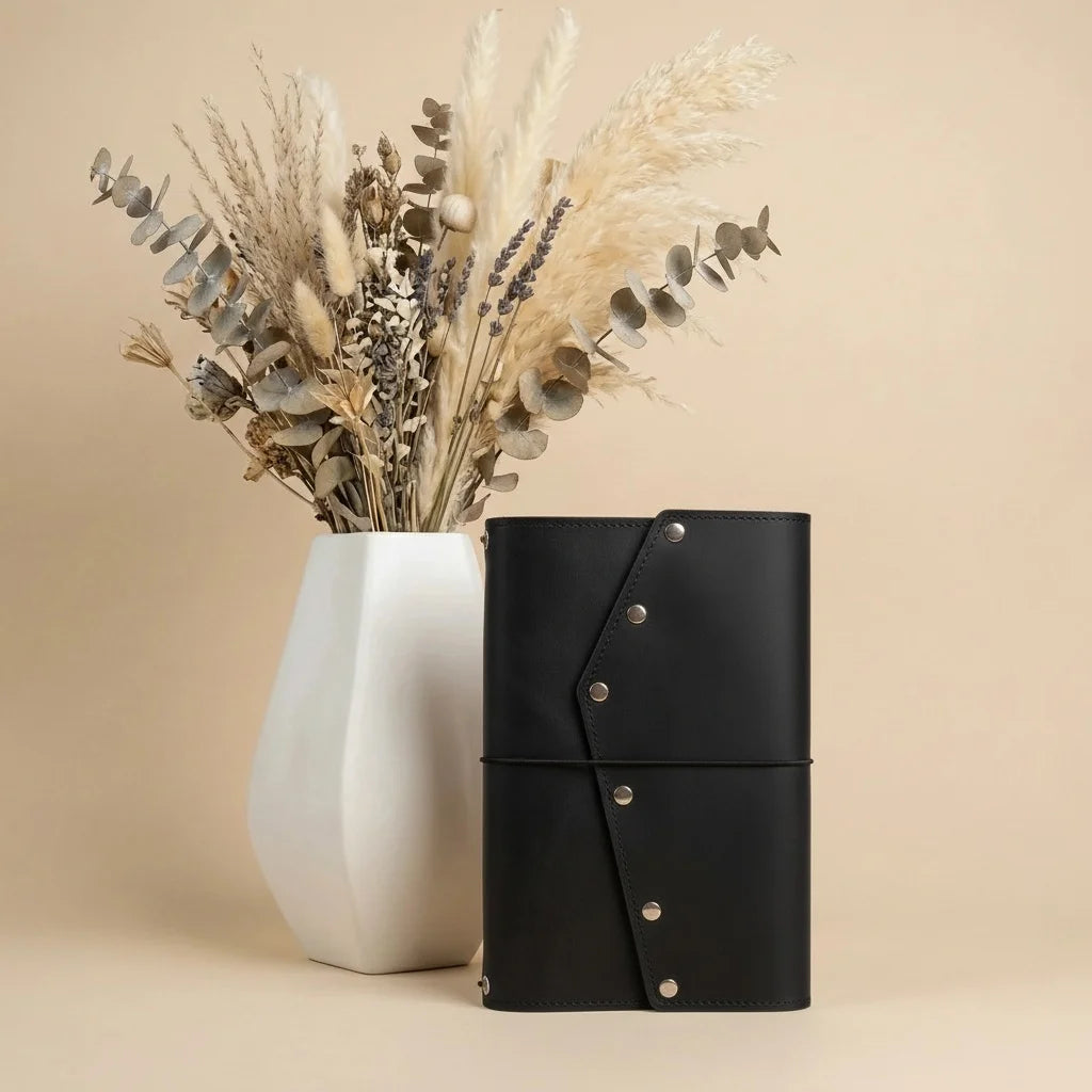 Black leather-bound notebook with silver buttons next to a white vase with dried plants on a beige background