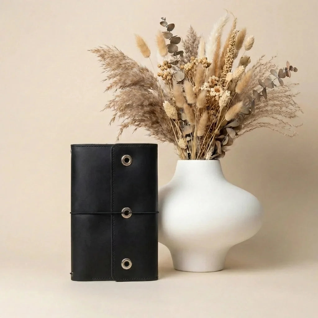 Black leather-bound book with a white vase containing dried plants on a beige background