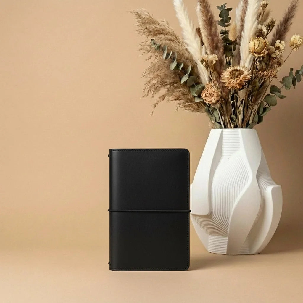 Brown leather notebook next to a white vase with dried plants on a beige background