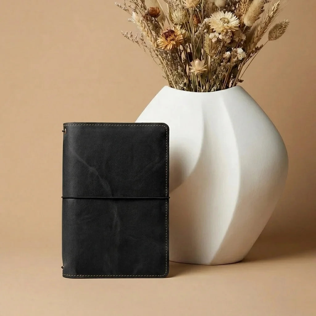 Black leather notebook next to a white vase with dried plants on a beige background