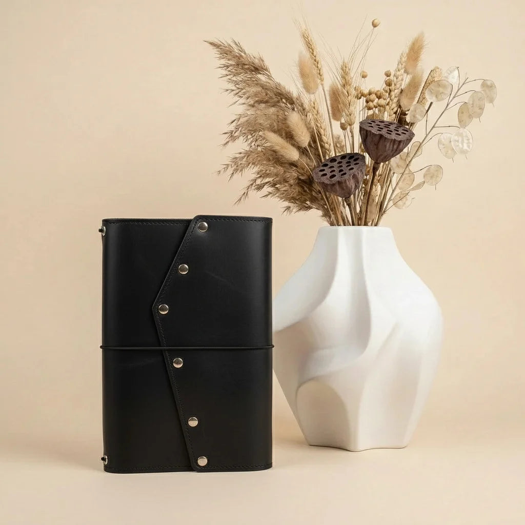 Black leather-bound book with silver buttons next to a white vase with dried plants on a beige background