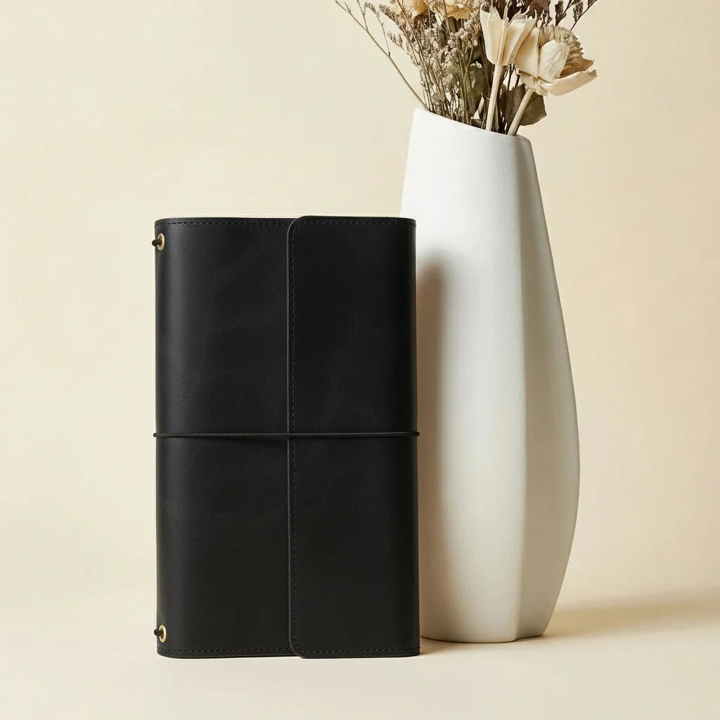 Black leather notebook next to a white vase with dried flowers on a beige background