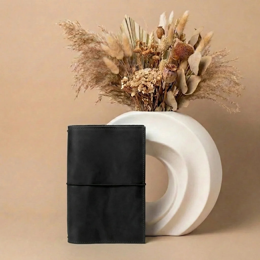 Black leather journal next to a white vase with dried flowers on a beige background