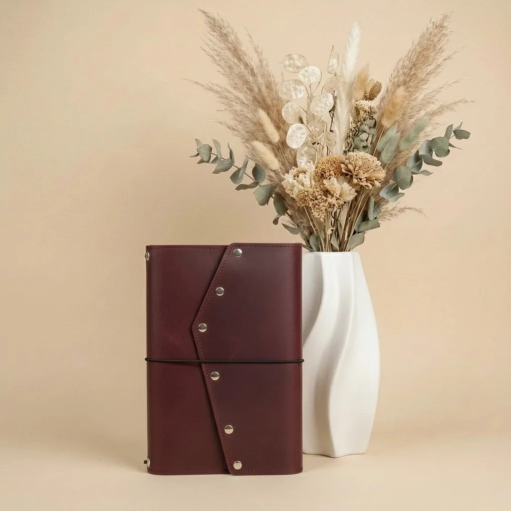 Brown leather notebook with silver buttons next to a white vase with dried flowers on a beige background