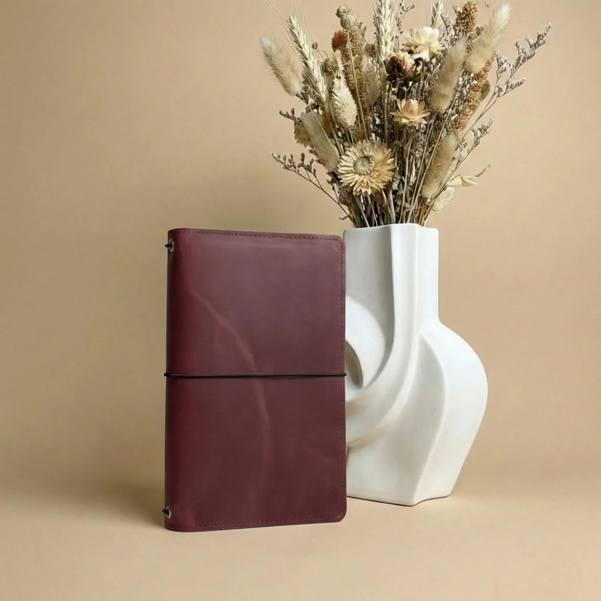 Maroon notebook next to a white vase with dried flowers on a beige background
