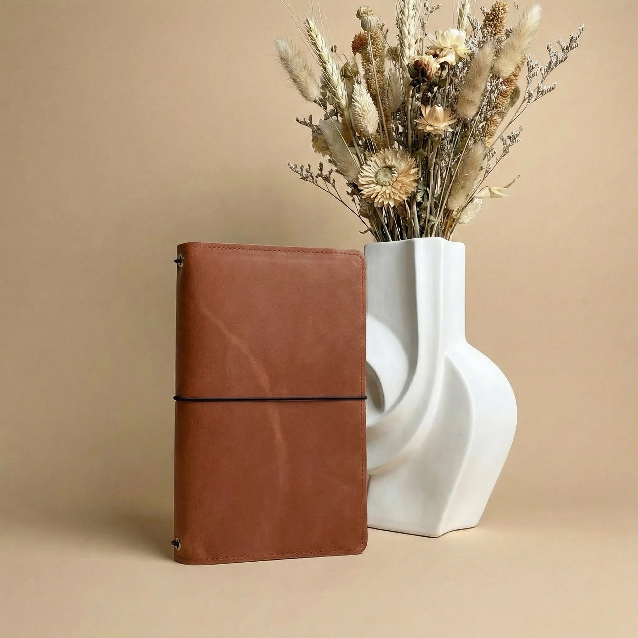 Brown leather notebook next to a white vase with dried flowers on a beige background