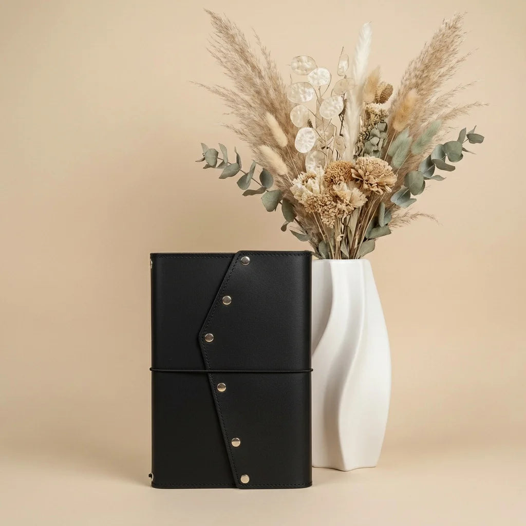 Black leather-bound notebook with gold buttons next to a white vase with dried flowers on a beige background