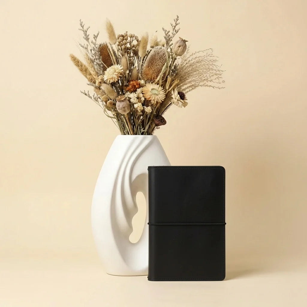Black notebook next to a white vase with dried flowers on a beige background