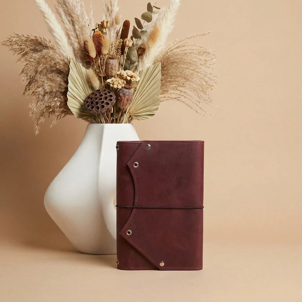Brown leather notebook with decorative elements next to a white vase with dried plants on a beige background