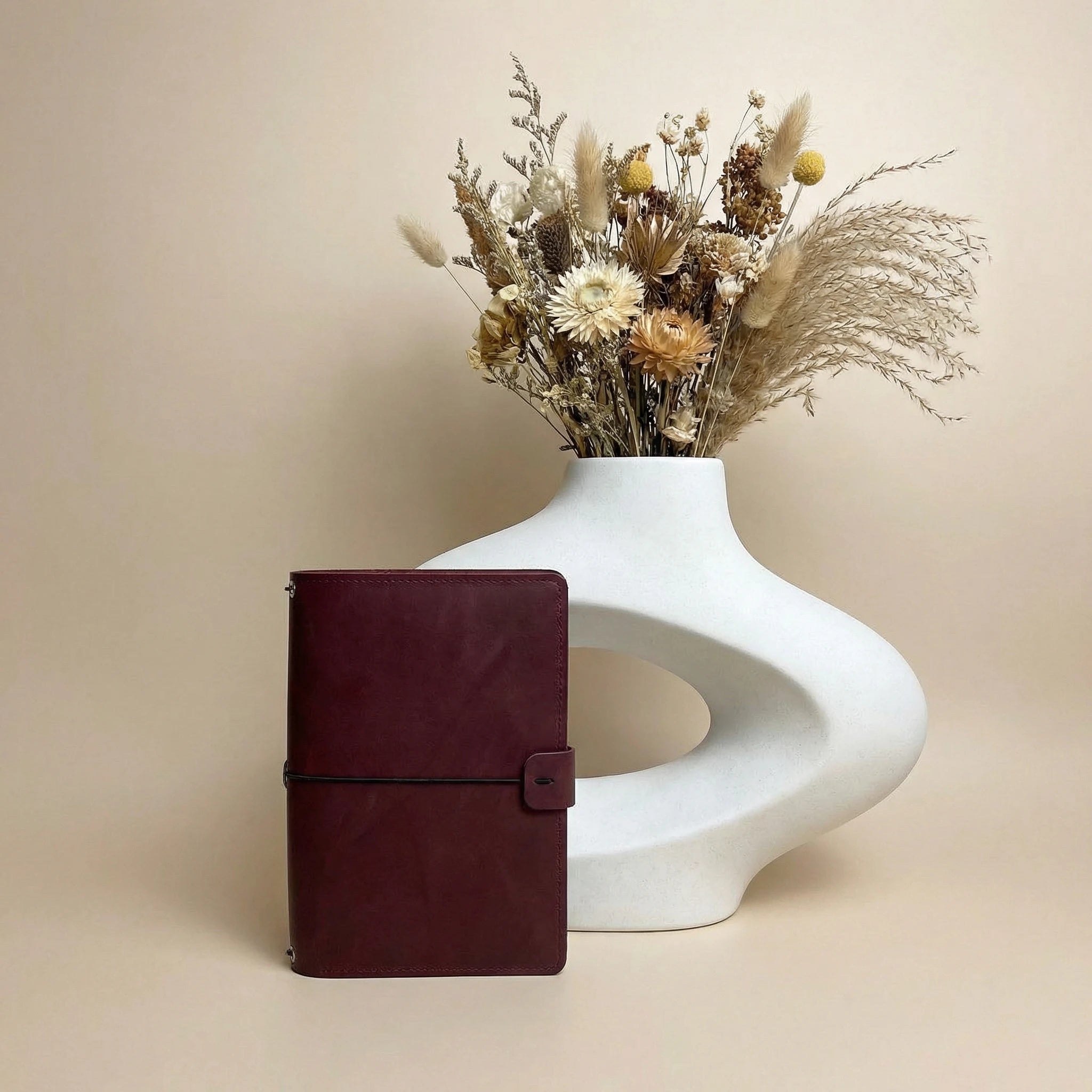 Maroon leather notebook next to a white vase with dried flowers on a beige background
