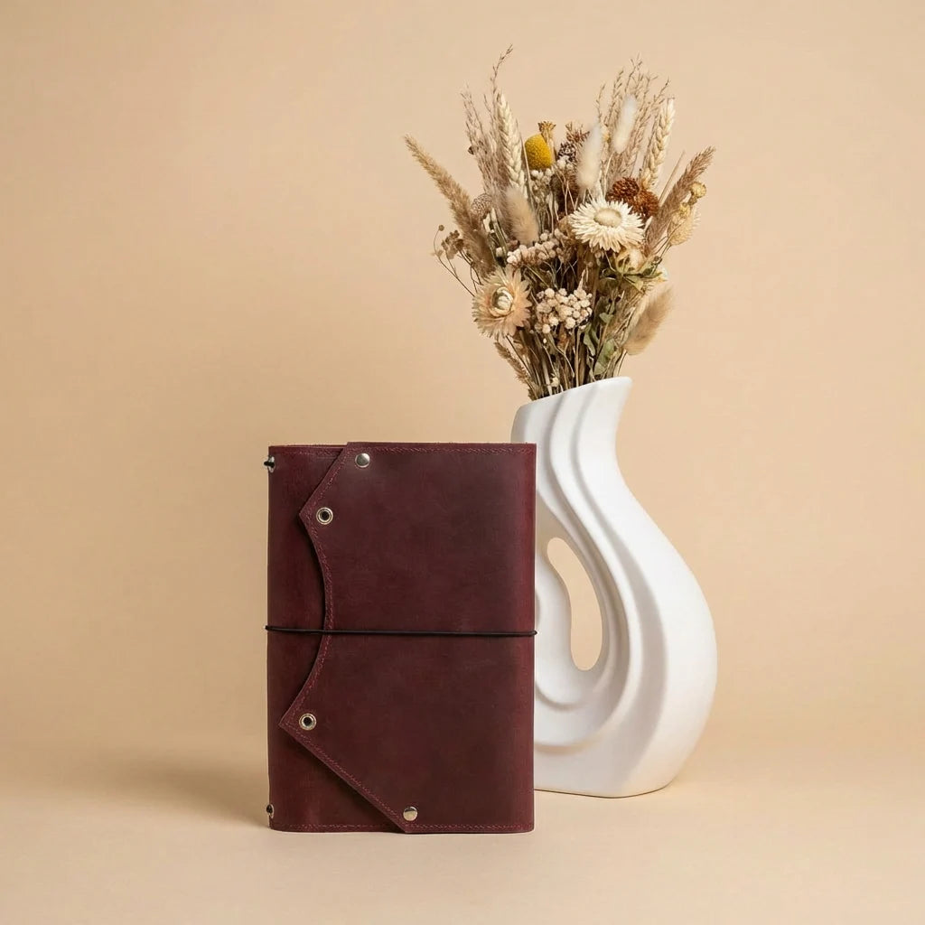 Brown leather notebook next to a white vase with dried flowers on a beige background