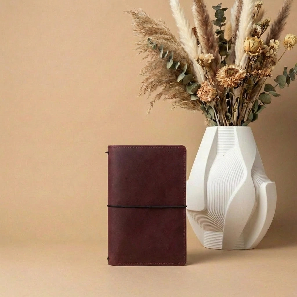Brown leather notebook next to a white vase with dried plants on a beige background