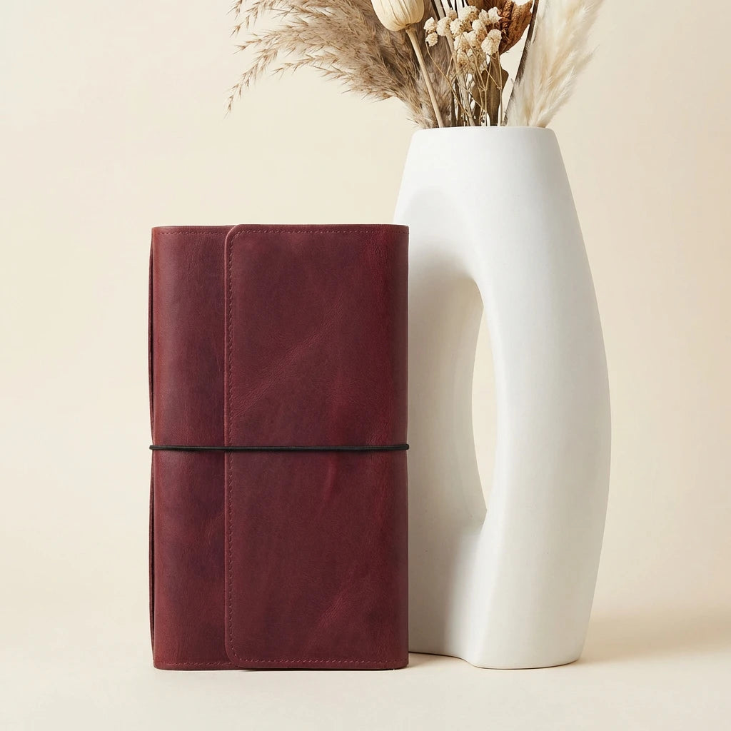 Maroon leather notebook next to a white vase with dried plants on a beige background