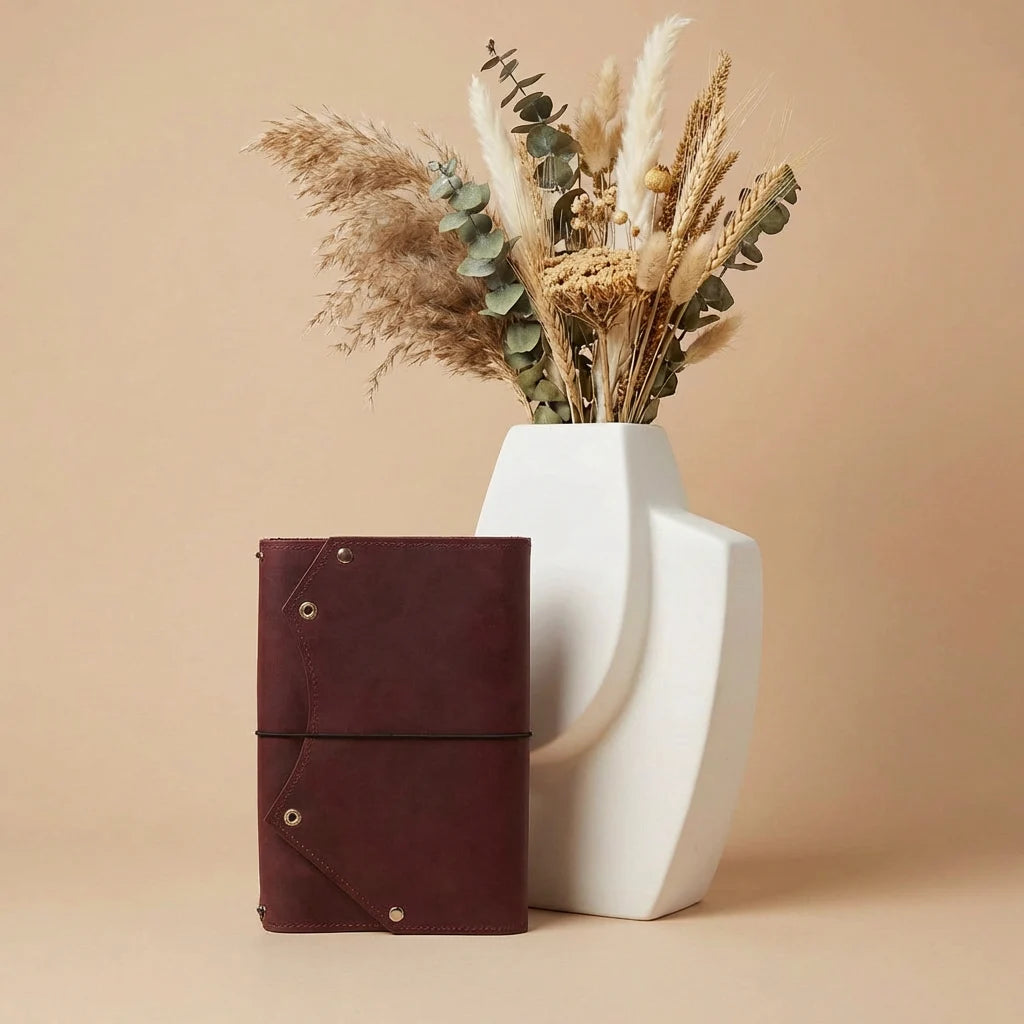 Brown leather notebook next to a white vase with dried plants on a beige background
