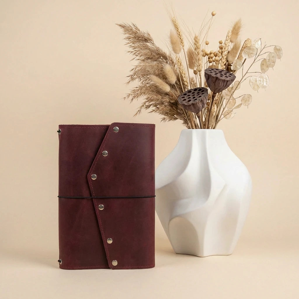 Brown leather notebook with silver buttons next to a white vase with dried plants on a beige background