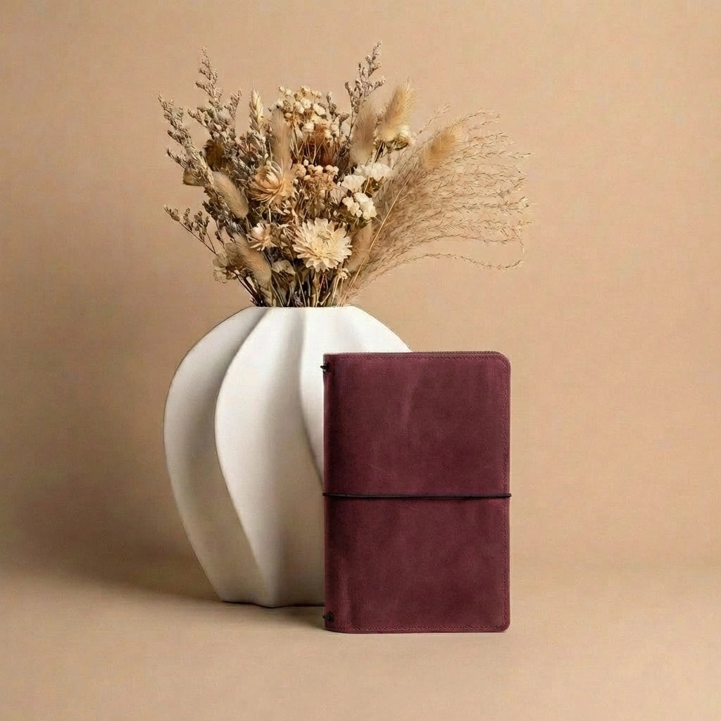 White vase with dried flowers next to a burgundy notebook on a beige background