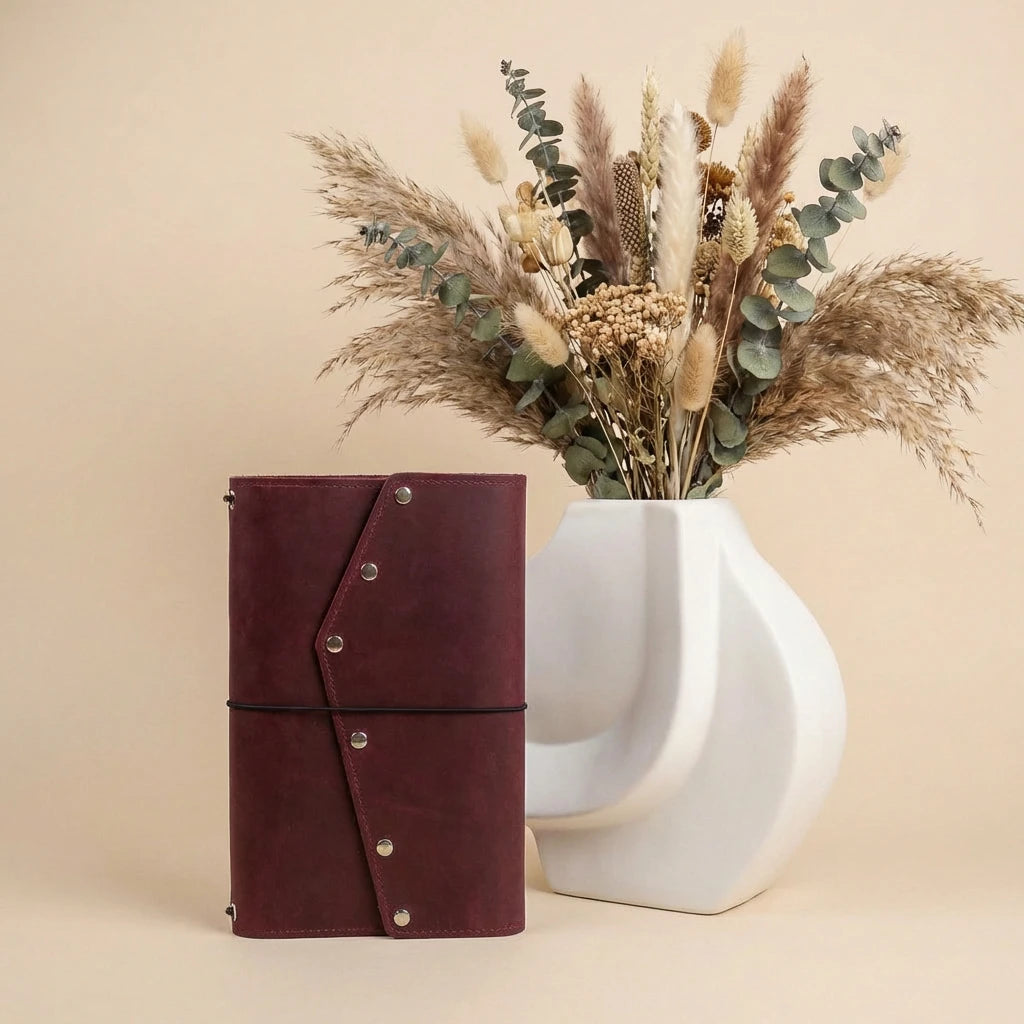 Brown leather notebook with silver buttons next to a white vase with dried flowers on a beige background
