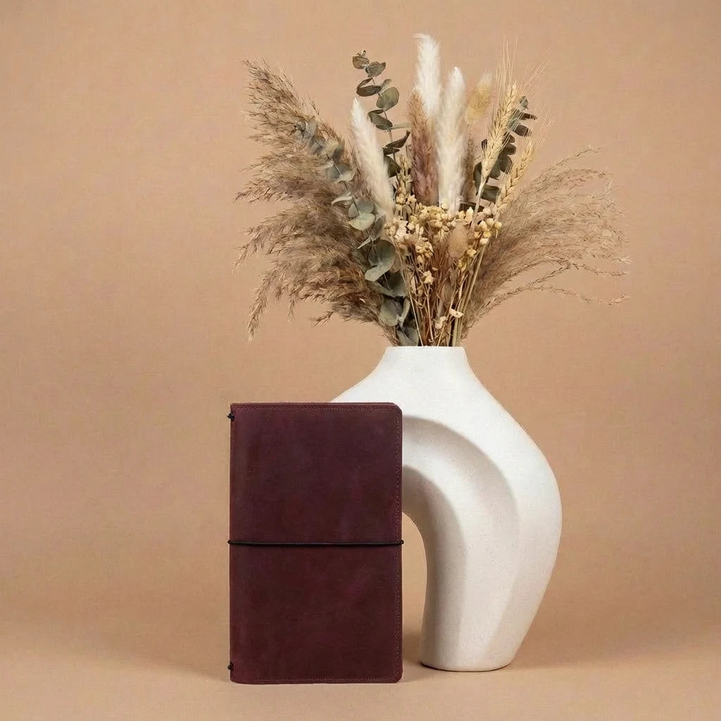 White vase with dried plants next to a brown leather notebook on a beige background