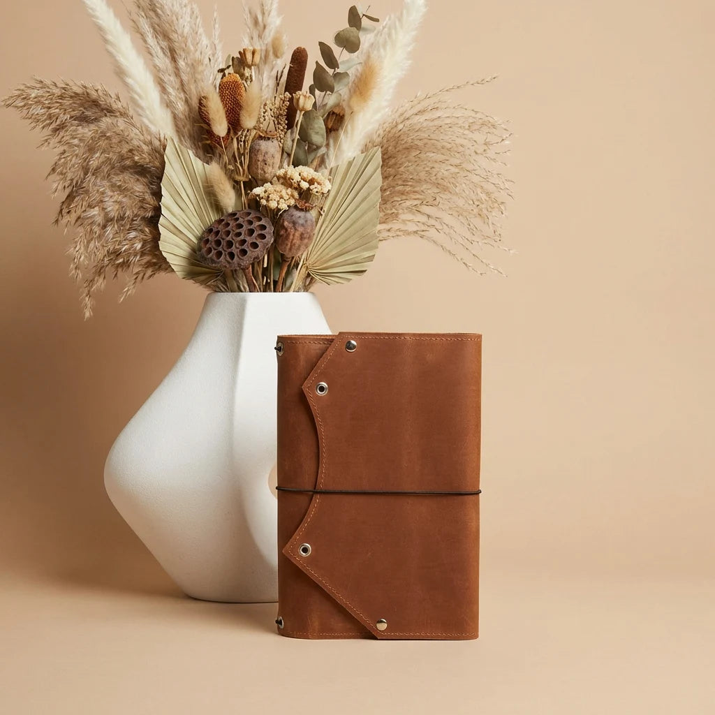 Brown leather notebook next to a white vase with dried plants on a beige background