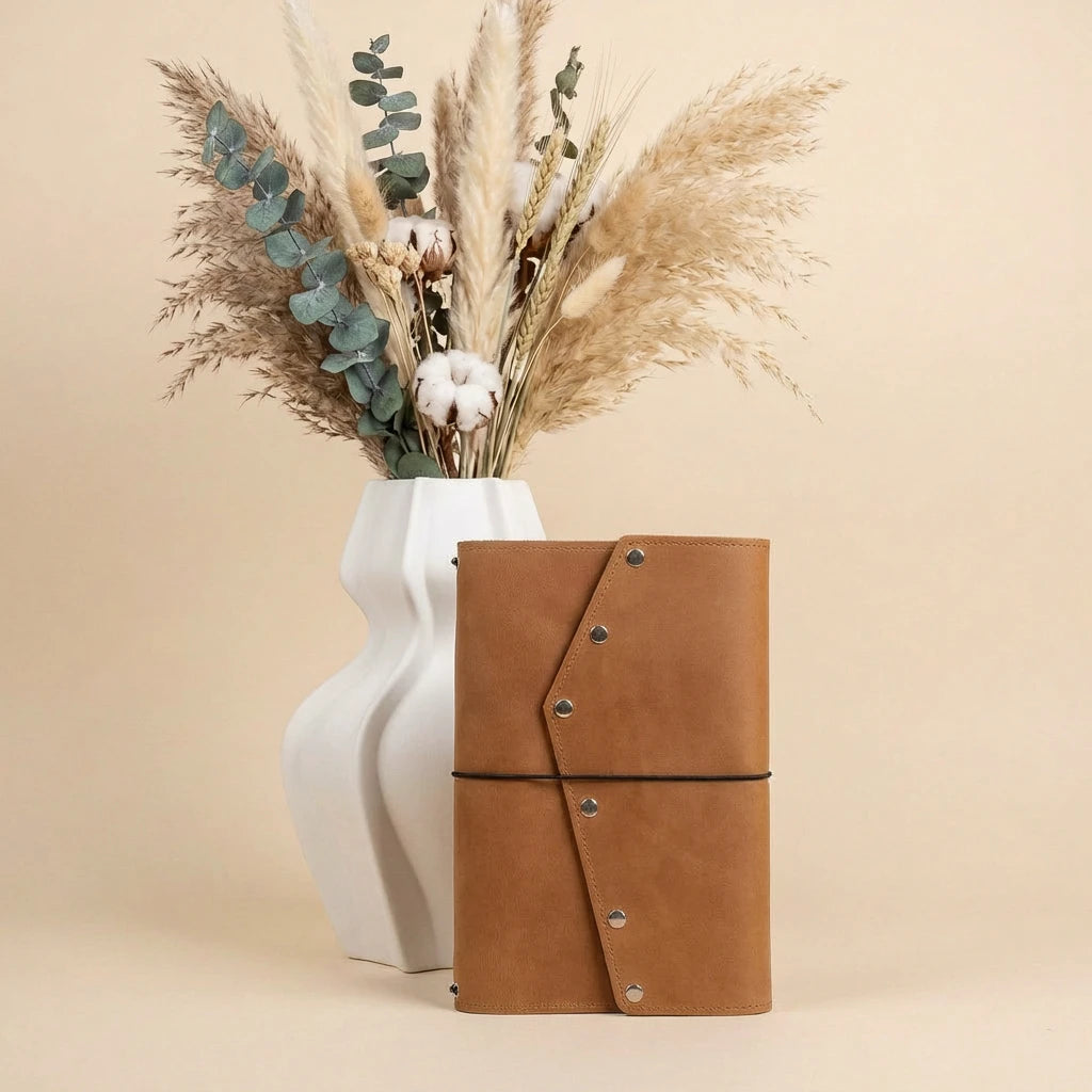 Brown leather notebook with decorative elements next to a white vase with dried plants on a beige background