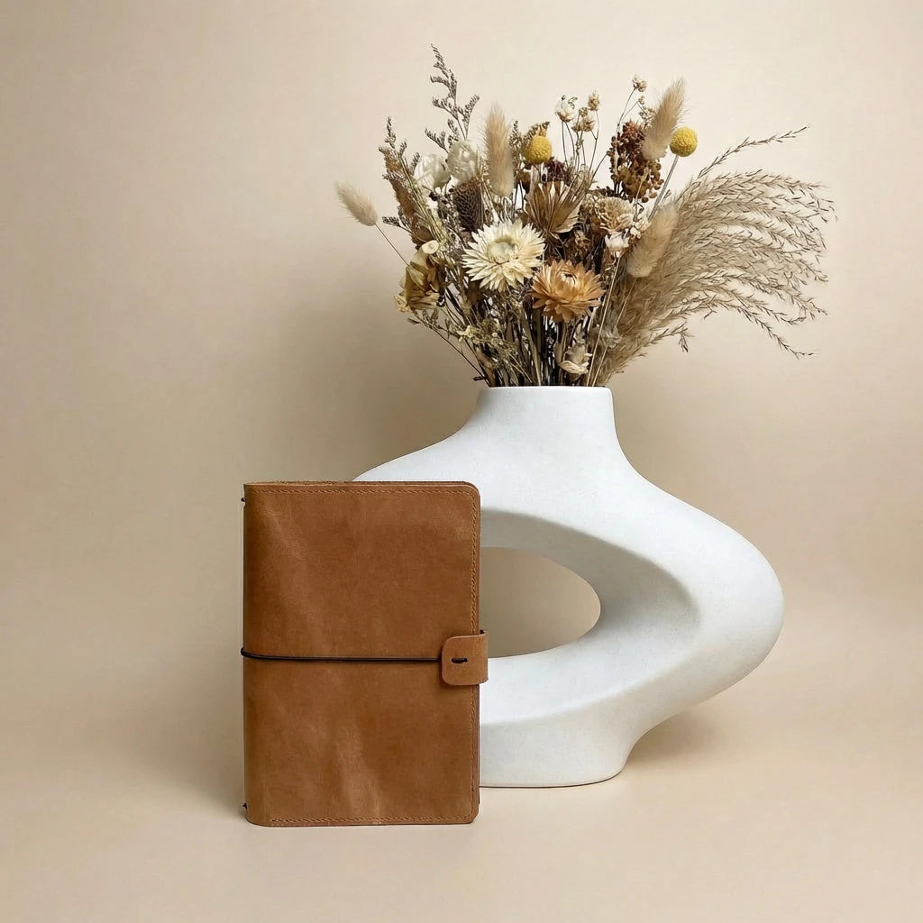 Brown leather notebook next to a white vase with dried flowers on a beige background