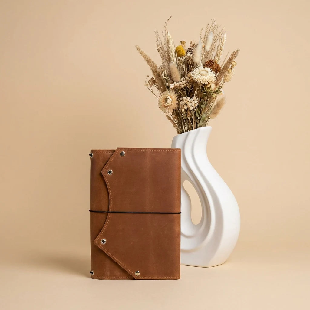 Brown leather notebook next to a white vase with dried flowers on a beige background
