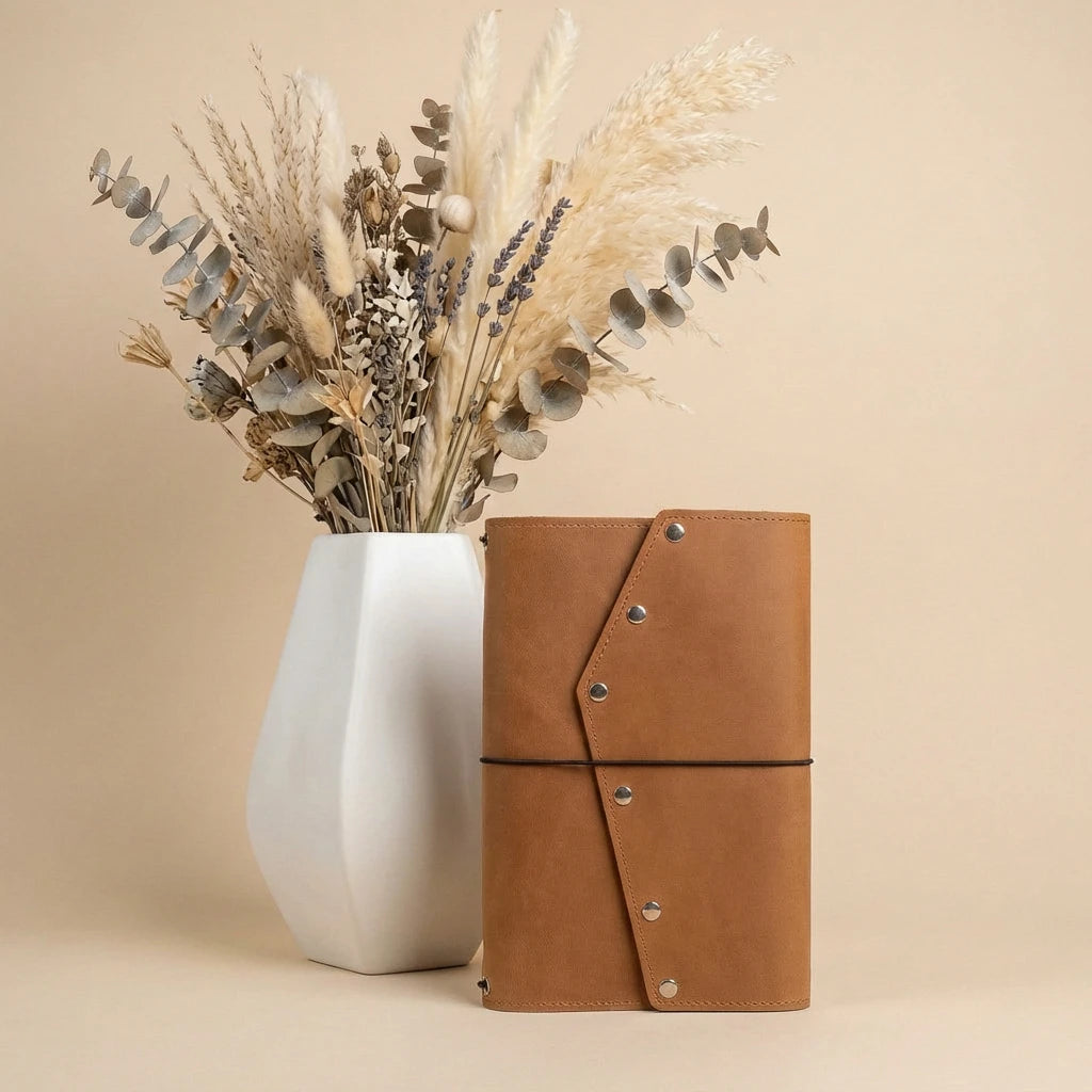 Brown leather notebook and white vase with dried plants on a beige background