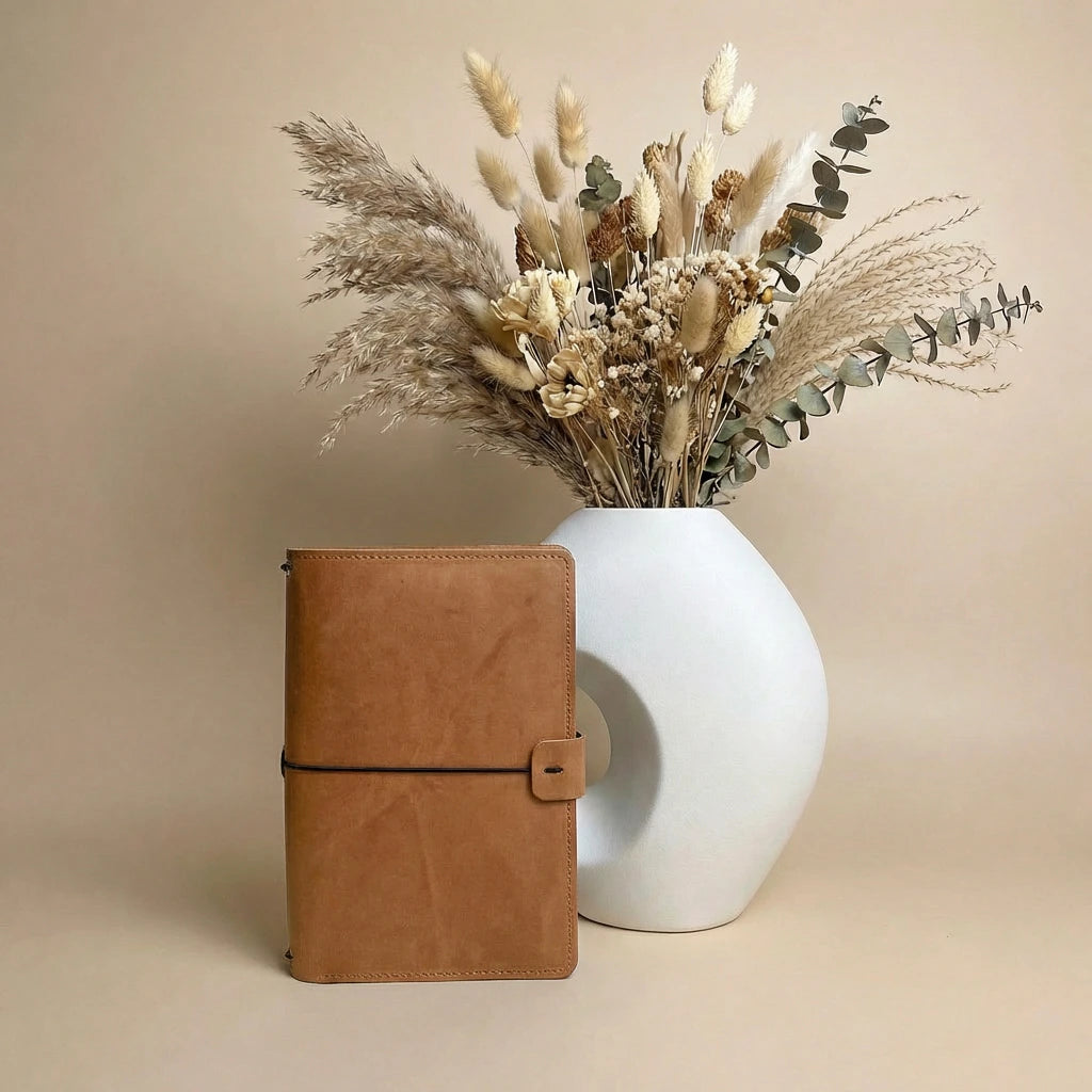 Brown leather notebook next to a white vase with dried flowers on a beige background