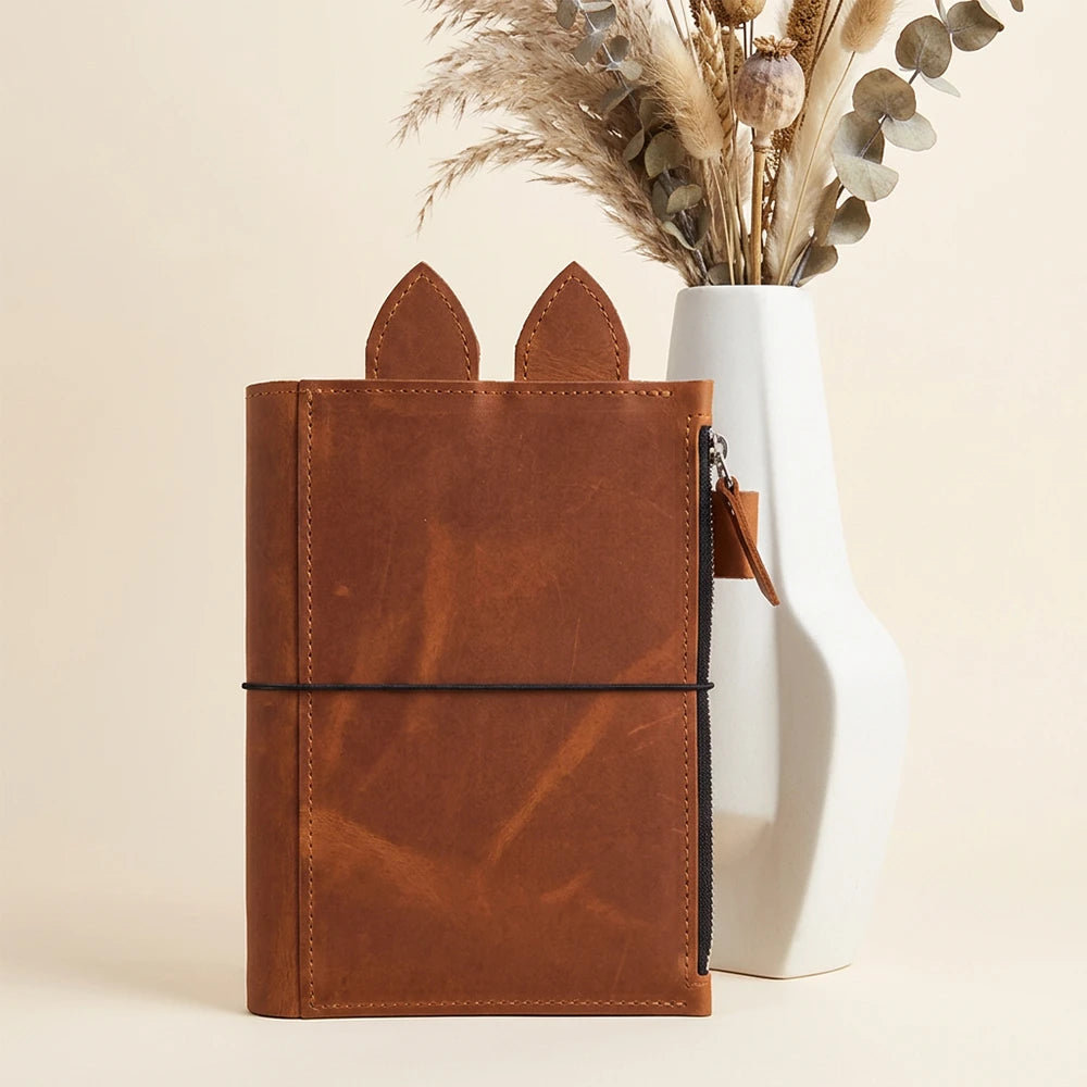 Refillable journal cover crafted from genuine leather with zipper pocket, pen loop, ears, and elastics.