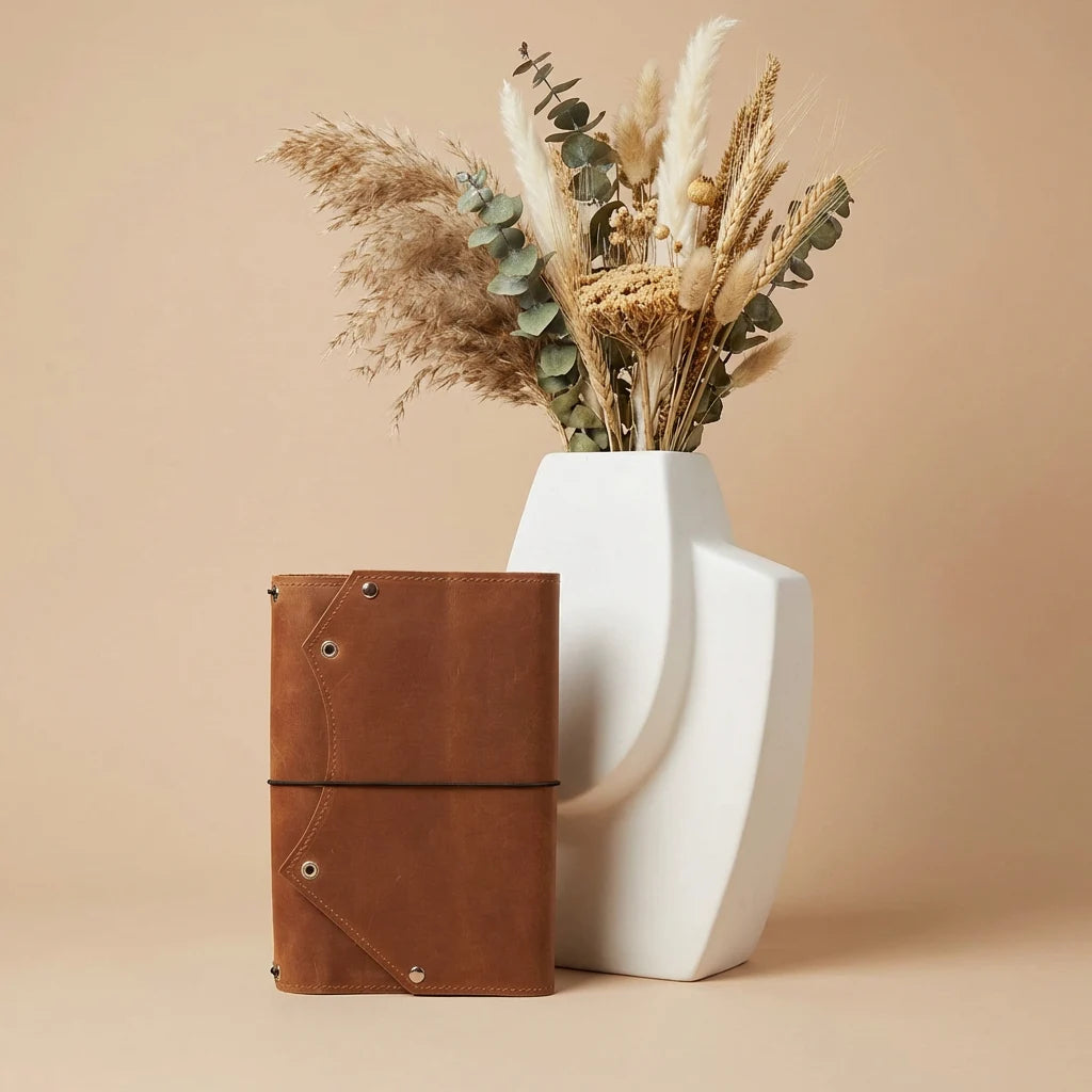 Brown leather notebook next to a white vase with dried plants on a beige background