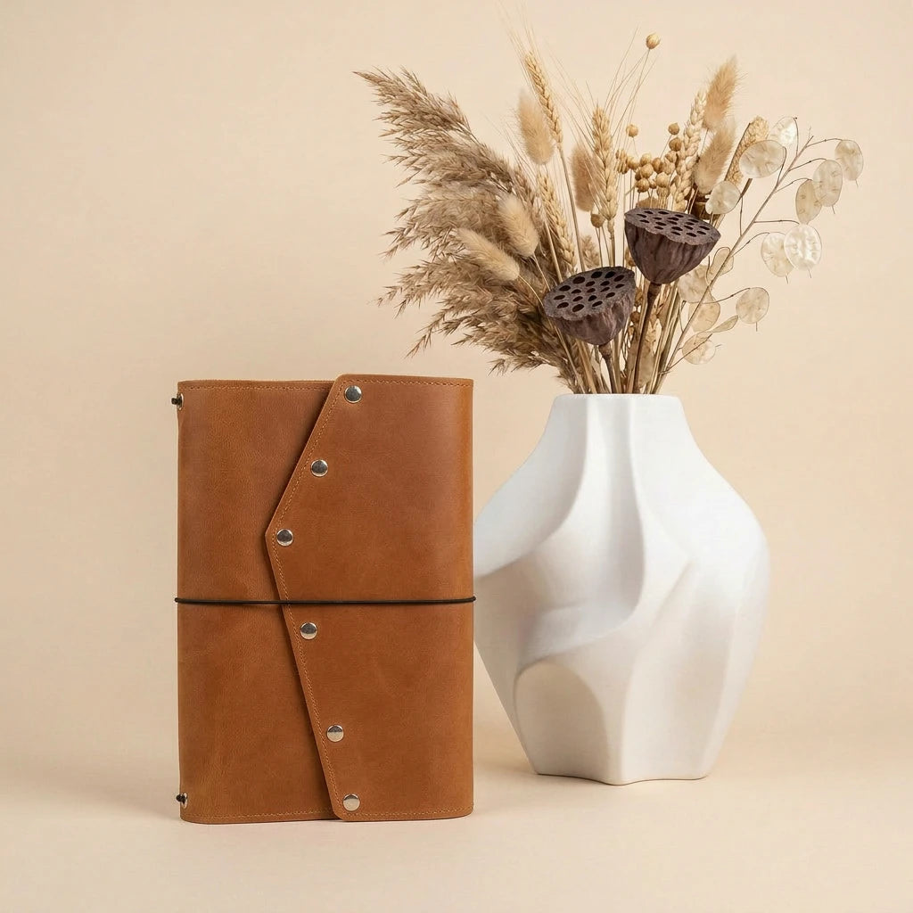 Brown leather notebook with a white vase containing dried plants on a beige background