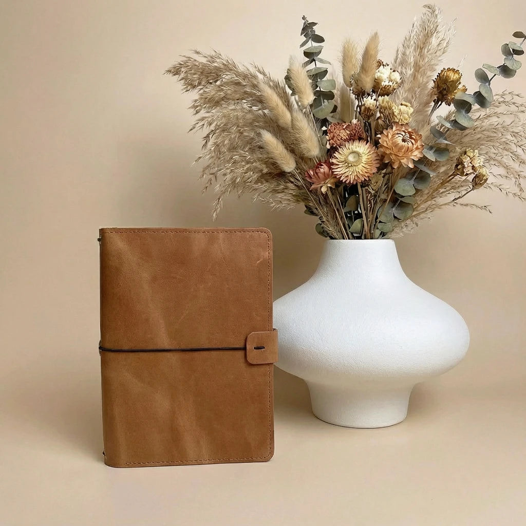 Brown leather notebook next to a white vase with dried flowers on a beige background
