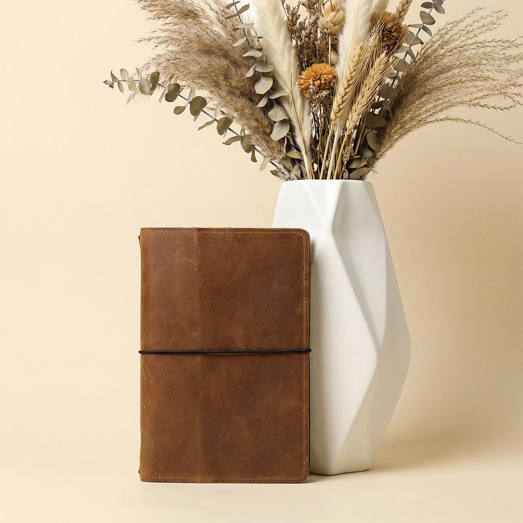 Brown leather notebook next to a white vase with dried plants on a beige background