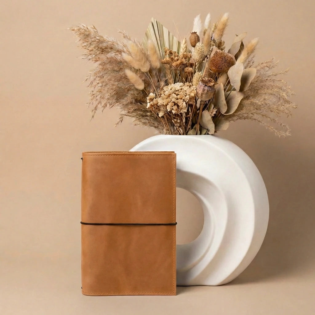 Brown leather notebook next to a white vase with dried flowers on a beige background