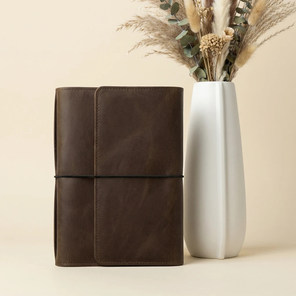 Brown leather notebook next to a white vase with dried plants on a beige background