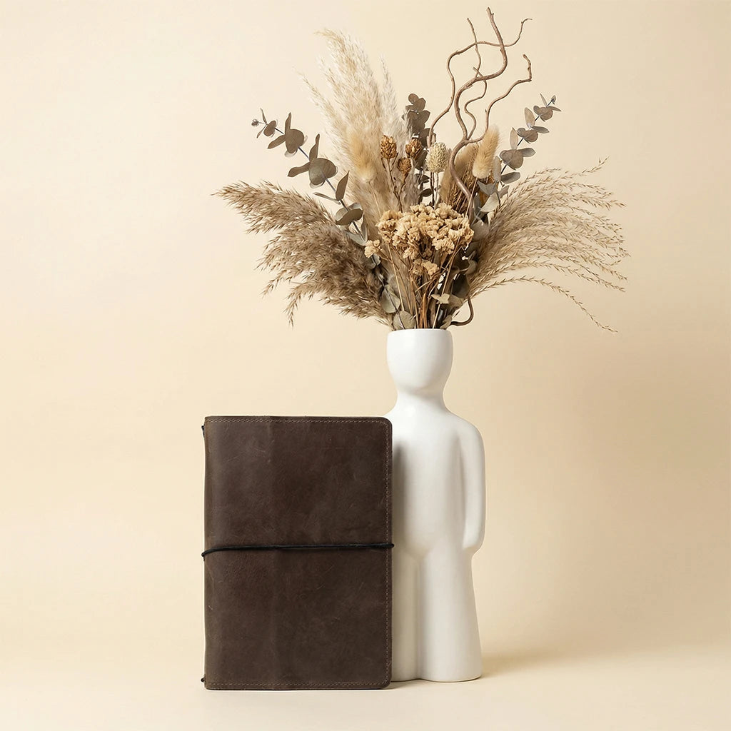 White vase with dried flowers next to a brown leather notebook on a beige background