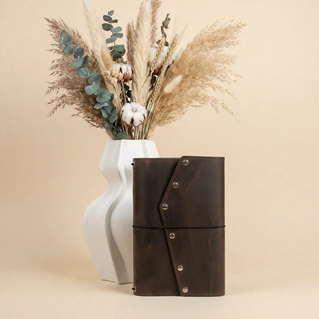 Brown leather notebook with decorative buttons next to a white vase with dried plants on a beige background
