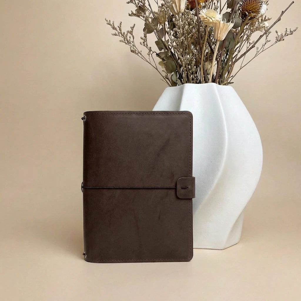Brown leather notebook next to a white vase with dried flowers on a beige background