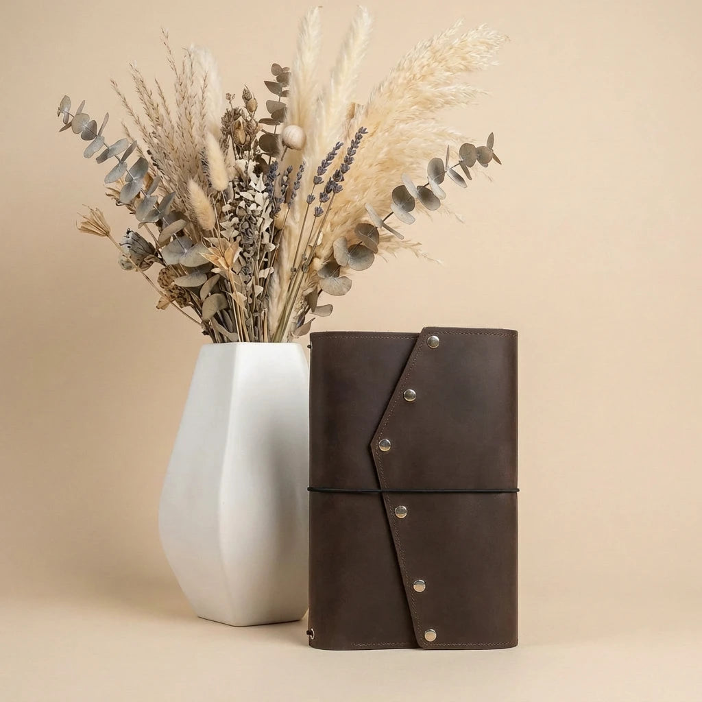 White vase with dried flowers next to a brown leather notebook on a beige background