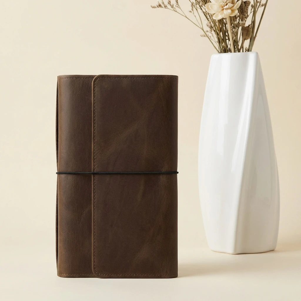 Brown leather notebook next to a white vase with dried flowers on a beige background