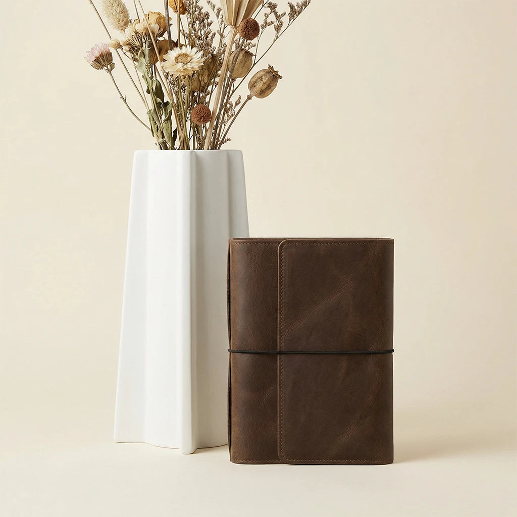 Brown leather notebook next to a white vase with dried flowers on a beige background