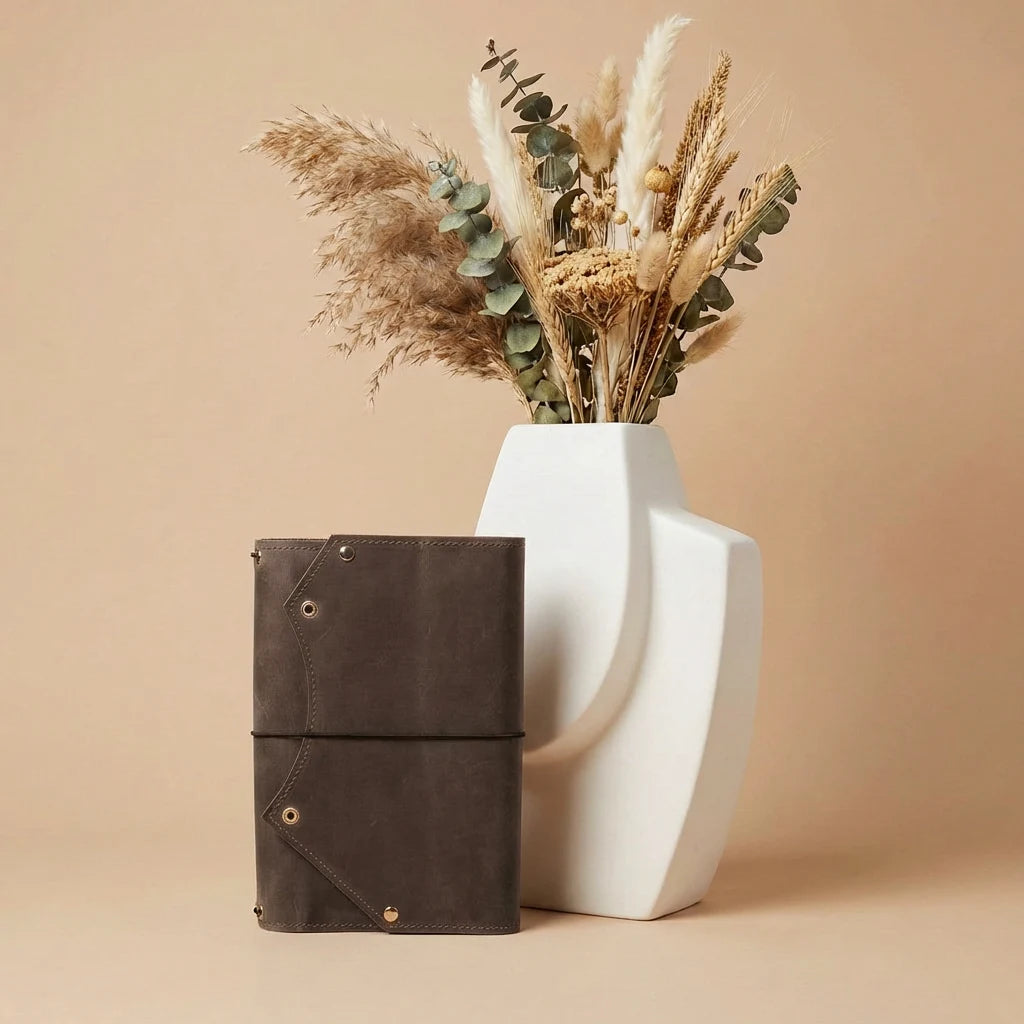 Brown leather notebook next to a white vase with dried plants on a beige background
