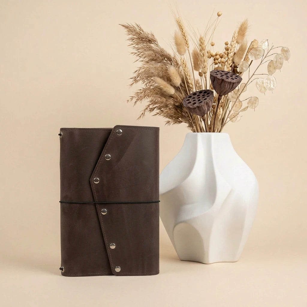 Brown leather notebook with silver buttons next to a white vase with dried flowers on a beige background