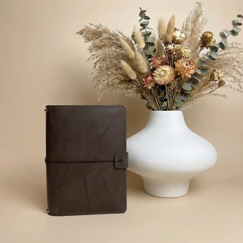 Brown leather notebook next to a white vase with dried flowers on a beige background