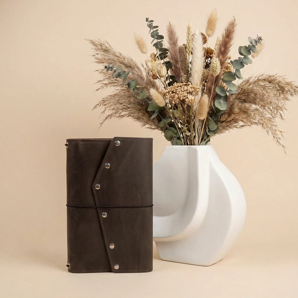Brown leather notebook with silver buttons next to a white vase with dried flowers on a beige background
