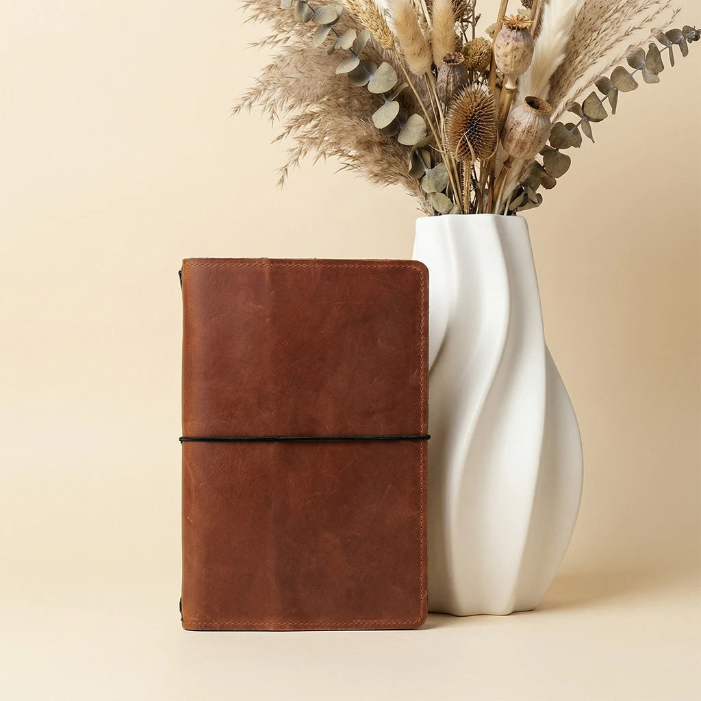 Brown leather notebook next to a white vase with dried flowers on a beige background