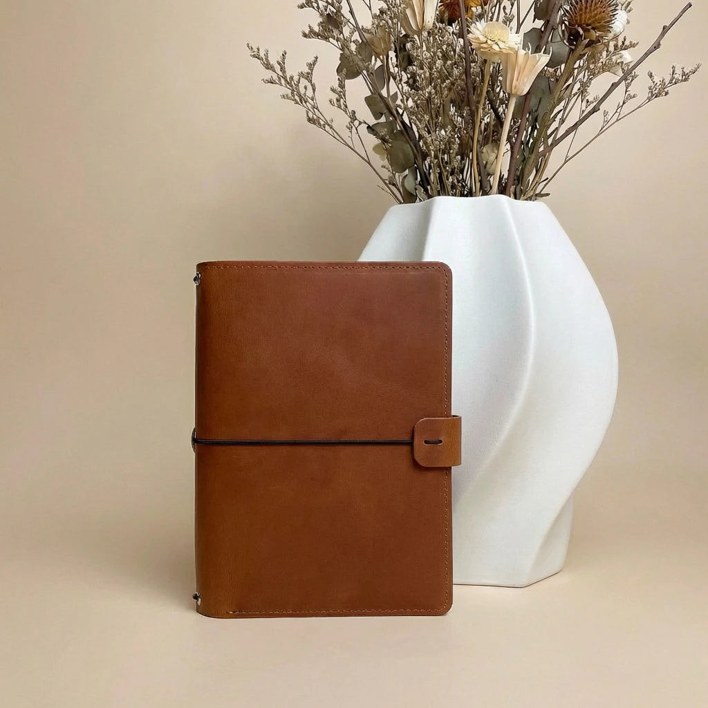 Brown leather notebook next to a white vase with dried flowers on a beige background