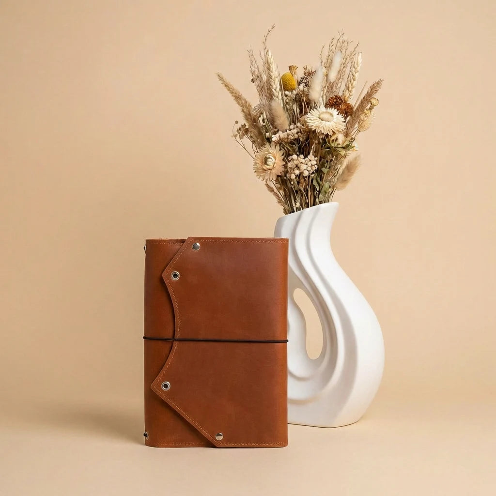 Brown leather notebook next to a white vase with dried flowers on a beige background