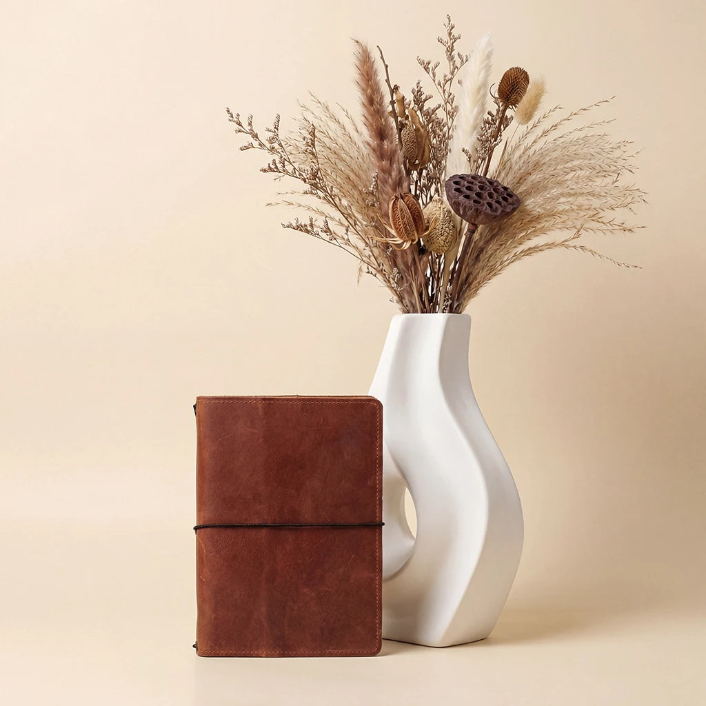 Brown leather notebook next to a white vase with dried flowers on a beige background